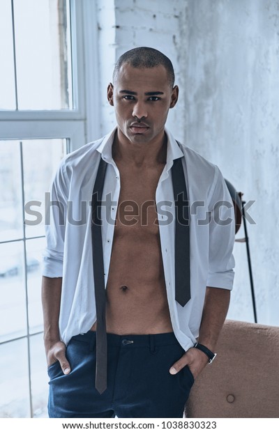 Real man.\
Handsome young African man in fully unbuttoned white shirt looking\
at camera while standing at\
home