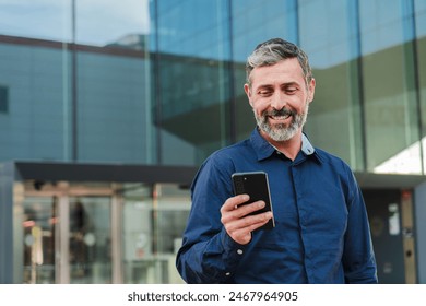 Real mature male worker watching a finance app on his cellphone. Middle aged person holding a mobile phone smiling. Handsome mid adult man smiling using a smartphone at workplace. High quality photo - Powered by Shutterstock