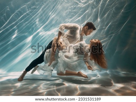 Real People art photo. Happy couple in love swim underwater, woman muse inspires male writer poet creator. Nymph girl dancing with guy at bottom sea under water. Red hair white long silk dress float.
