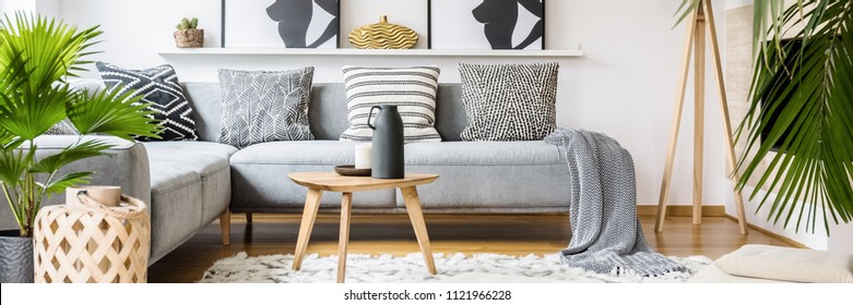 Real photo of black jug and white candle placed on small wooden table standing on fluffy carpet in bright living room interior with corner grey couch and fresh plants