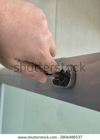 Real photo capturing a close-up angle of a human hand holding and inserting a metal key into a stainless steel cylinder lock mounted on the patch fitting frame of a glass door.