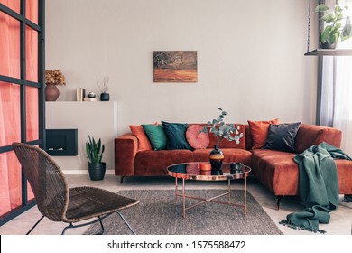 Real Photo Of A Comfy Living Room Interior With A Round Table On Gray Rug, Wicker Armchair And Red Corner Sofa