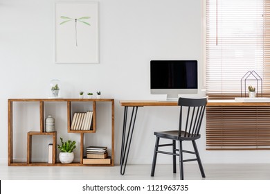 Real photo of a computer with empty screen standing on a wooden desk in bright freelancer's office interior with black chair and bookcase