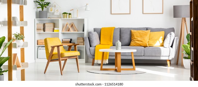 Real Photo Of A Couch With Yellow Pillows Standing Next To Shelves With Books, Ornaments And Lamp. Table And Armchair In Spacious Living Room Interior With Posters On The Wall