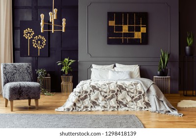 Real Photo Of Fresh Plants And Black And Gold Paintings Hanging On Walls With Molding In Dark Bedroom Interior With King-size Bed And Floral Armchair