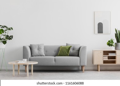 Real photo of grey lounge with green cushion, wooden coffee table, simple poster on the wall and cupboard with books in bright sitting room interior - Shutterstock ID 1161606088