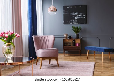 Real Photo Of A Pink Armchair Standing On A Rug And Under A Lamp In Spacious Living Room Interior, Next To A Table With Flowers And In Front Of A Shelf Next To A Grey Wall With Dark Painting