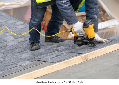 Real Photo Of Professional Roofer Worker In Uniform Work Wear Using Air Or Pneumatic Nail Gun And Installing Asphalt Or Bitumen Shingle On Top Of The New Roof Under Construction Residential Building