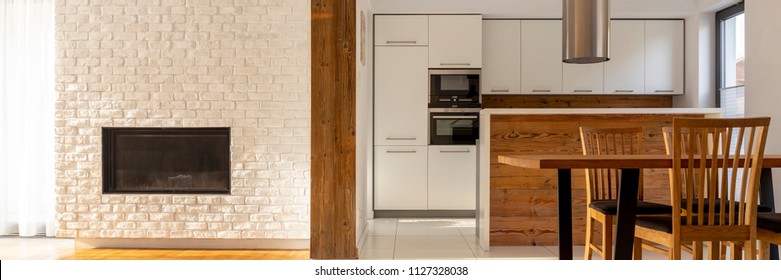 Real photo of white open space kitchen interior with two ovens, wooden countertop and dining table with chairs. Empty place on the wall above fireplace for your TV