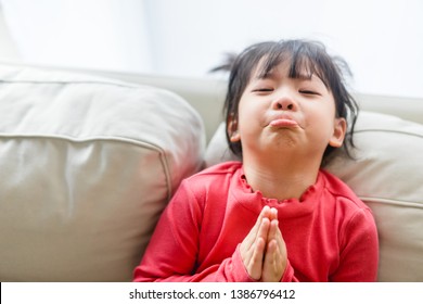 Real shot of regretful kid girl looks gloomy and desperate, keeps palms together, beggs for forgiveness in her mother at home, feels sorry and guilty. Cute adorable asian girl pleads about something.