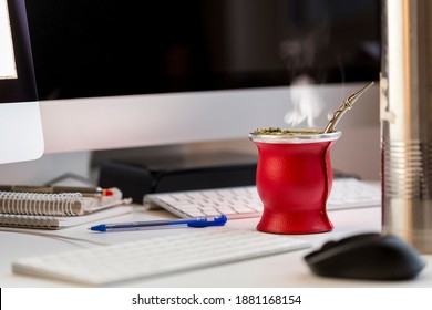 Real Steam In Hot South American, Caffeine-Rich Infused Drink Yerba Mate In Red Gourd In Editor's Modern Office Environment Workplace With Computer Equipment, Keyboards, Mouse, Hard Disk, Notepads.