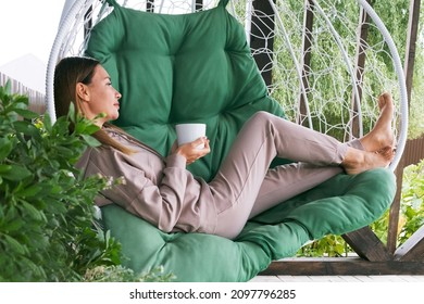 Real Woman Drinking Tea Or Coffee Outdoor. Full Length Mature Woman Sitting In A Hammock Chair And Relaxing In Nature. Woman Chilling Enjoy Relaxing 