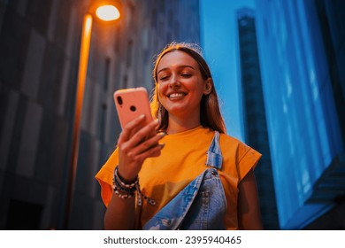 Real young adult woman using a cellphone at night. Happy female having fun with a smartphone social media app at dusk. Teenage girl browsing on internet with a mobile phone application at evening - Powered by Shutterstock