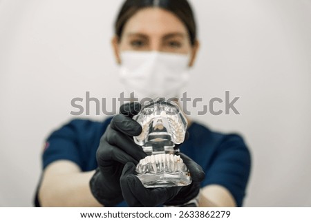 A realistic dental prosthesis is held up to the camera by a dentist, who appears out of focus in the background, highlighting the dental model in a clinical setting.