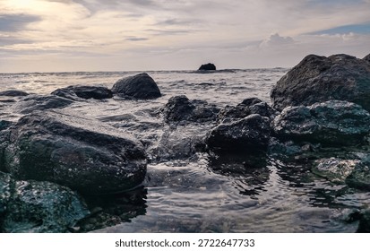 Realistic low-angle view of ocean waves washing over dark coastal rocks during a calm afternoon. Soft lighting, natural water textures, and serene seascape atmosphere.