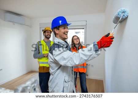 A realistic scene of an interior renovation in progress. In the foreground, a painter in a high-visibility vest and safety helmet is focused on painting a large white wall. Standing nearby, a group of construction workers and engineers.