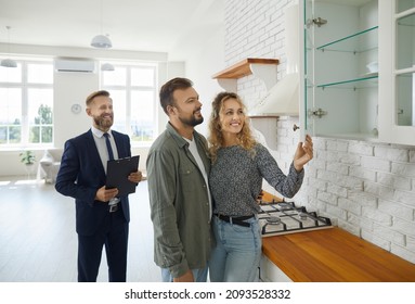 Realtor With Customers Checking Apartmnet. Couple Examines Furniture In Kitchen While Inspecting House With Real Estate Agent. Friendly Male Realtor Advises Young Family Before Buying New Home.