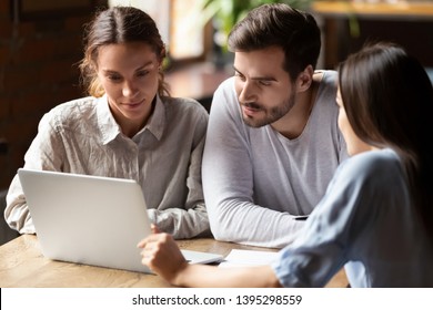 Realtor Or Insurance Broker Consulting Young Couple In Cafe, Sitting Together At Table, Using Laptop, Looking At Screen, Financial Advisor Discussing Mortgage Or Loan Conditions With Clients