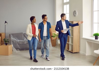 Realtor Or Real Estate Agent Giving House Tour To Future Buyers. Young Black Couple Planning To Buy New Home. Boyfriend And Girlfriend Or Husband And Wife Looking At Modern, Big, Spacious Apartment