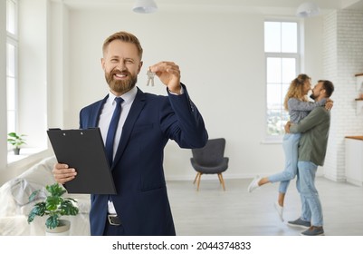 Realtor or real estate agent shows key to house he has sold. Portrait of man in suit standing in living room with happy married couple in background, holding folder and key, smiling, looking at camera - Shutterstock ID 2044374833