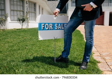 Realtor With A For Sale Sign Board Outside A House. Male Real Estate Broker With For Sale Notice In Lawn Of A New House.