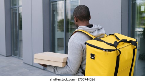 Rear Of African American Young Man Walking The Street With Big Yellow Backpack While Working As Deliveryman. Delivering Of Food Concept By Walk. Back View On Guy Carrying Pizza Boxes.