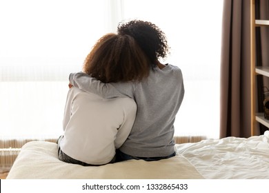 Rear back view black mother and daughter embrace sitting on bed at home, older sister consoling younger teen, girl suffers from unrequited love share secrets trustworthy person relative people concept