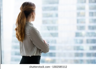 Rear back view at confident rich businesswoman looking forward through glass window of skyscraper building dreaming of success thinking of future business vision and new goals enjoying city view