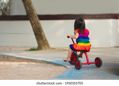Rear back view of cute girl riding red tricycle on playground blue road. Funny kid recreation. Active child wearing colorful dress. Along the road there is a sand pit. Happy children aged 4 years old.