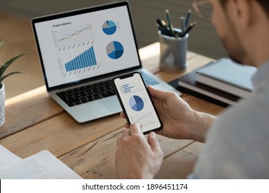Rear back view focused young businessman analyzing marketing research results on smartphone and computer, developing growth strategy, checking sales data statistics using modern gadgets in office.