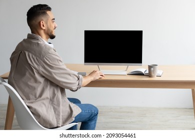 Rear back view of happy Middle Eastern man looking at blank empty computer monitor sitting at desk at workplace, watching webinar or having online web video call, browsing internet, free copy space