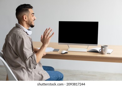 Rear Back View Of Happy Middle Eastern Man Having Online Web Video Call, Looking And Waving Hand At Blank Empty Computer Monitor Webcam Sitting At Desk At Workplace, Free Copy Space Mock Up