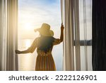 Rear back view woman opening curtains, relaxing and looking beautiful seascape panorama with rays of sun light in holiday on the balcony in high quality hotel feeling happy. Composition of nature