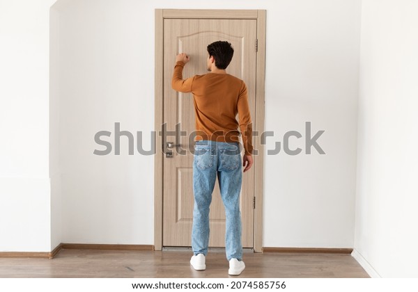 Rear back view of young\
casual man knocking on the wooden door, casual male visitor\
standing in entrance, guest wants to come in, full body length,\
free copy space