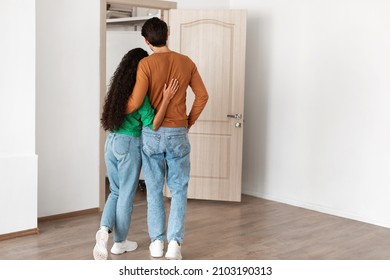 Rear Back View Of Young Couple Hugging Looking At Open Door Together, Welcoming Inviting Guests To Enter Home, Guy And Lady Standing In Doorway Of Modern Flat, Waiting For Visitor Friends To Come In