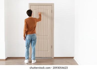 Rear back view of young man knocking on the wooden closed door, male visitor standing in front entrance, guest wants to come in, free copy space, full body length