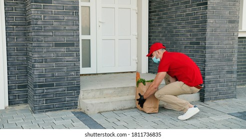 Rear Of Caucasian Male Courier In Medical Mask Delivering Bag With Food To Client House And Leaving At Door. Delivery Man Bringing Packet To Customer Home. Shopping. Lockdown. Coronavirus Quarantine.