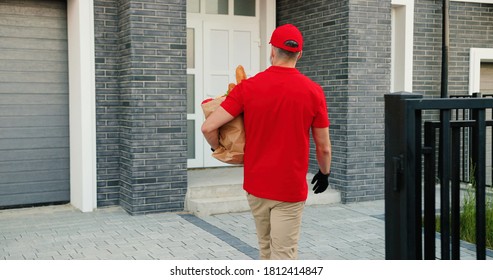 Rear Of Caucasian Male Courier In Medical Mask Delivering Bag With Food To Client House And Leaving At Door. Delivery Man Bringing Packet To Customer Home. Shopping. Lockdown. Coronavirus Quarantine.