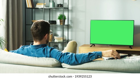 Rear of Caucasian young guy watching TV with chroma key resting on sofa sitting in living room in house. Man watch film on television with green screen sitting on couch in apartment, leisure
