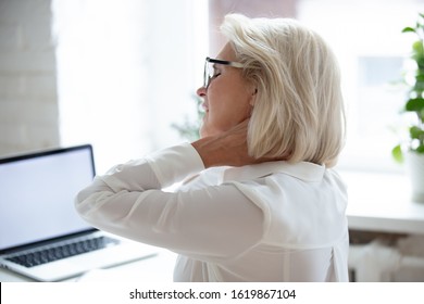 Rear close up view at workplace tired middle-aged business woman suffering from neck pain sitting at computer caused by long time laptop usage. Incorrect posture or desk setup, pinched nerve concept - Powered by Shutterstock