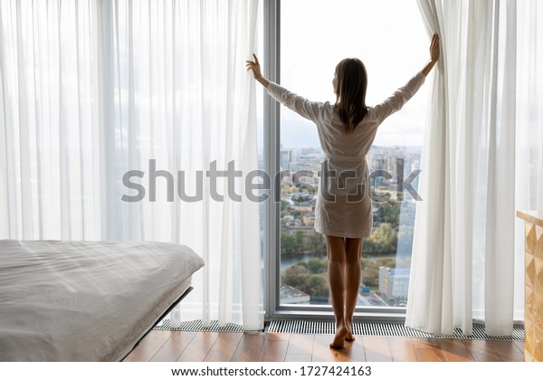 Rear full length of woman opened curtains enjoys\
big picturesque city standing barefoot on warm heated floor near\
panoramic window enjoy urban view, climate control at home,\
welcoming new day concept