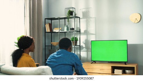 Rear Of Happy African American Couple Man And Woman Sitting On Couch In Living Room Watching At TV Monitor With Green Screen In Positive Mood. Family Cheering For Fav Team, Watching TV, Sport Match