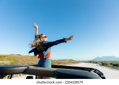 Rear High Angle View Of A Caucasian Woman Inside An Open Top Car, Standing, Smiling And Raising Her Hands. Weekend Beach Vacation, Lifestyle And Leisure.