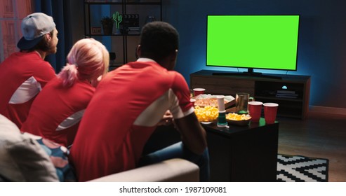 Rear Of Mixed-race Young Happy Male And Female Friends Sitting On Sofa In Dark Living Room At Home In Evening Watching TV With Green Screen Fans Cheering For Favorite Sport Team Entertainment Concept