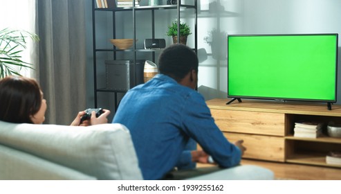 Rear of mixed-races couple African American man and Caucasian woman sitting on sofa in room watching at TV with green screen playing video games on console. Friends play game having fun, chroma key