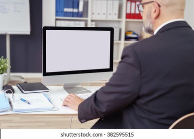 Rear Over The Shoulder View Of A Businessman Working At His Desk In The Office With Focus To His Blank Desktop Computer Monitor