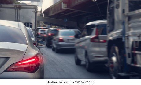 Rear perspective view of multiple vehicles queuing in traffic near an elevated highway structure. Bright daylight illuminates the urban commuting scene. Heavy traffic during rush hour.