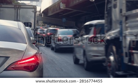 Rear perspective view of multiple vehicles queuing in traffic near an elevated highway structure. Bright daylight illuminates the urban commuting scene. Heavy traffic during rush hour.