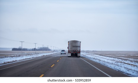 Rear of semi truck and trailer lined with warning lights driving down a highway in snowy weather in rural agricultural landscape