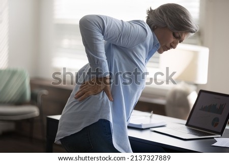 Rear side view aged woman stand near workplace desk, touch lower back, suffer from sudden backache, due to uncomfortable chair. Sedentary work disease, rheumatism, low-back lumbar pain illness concept